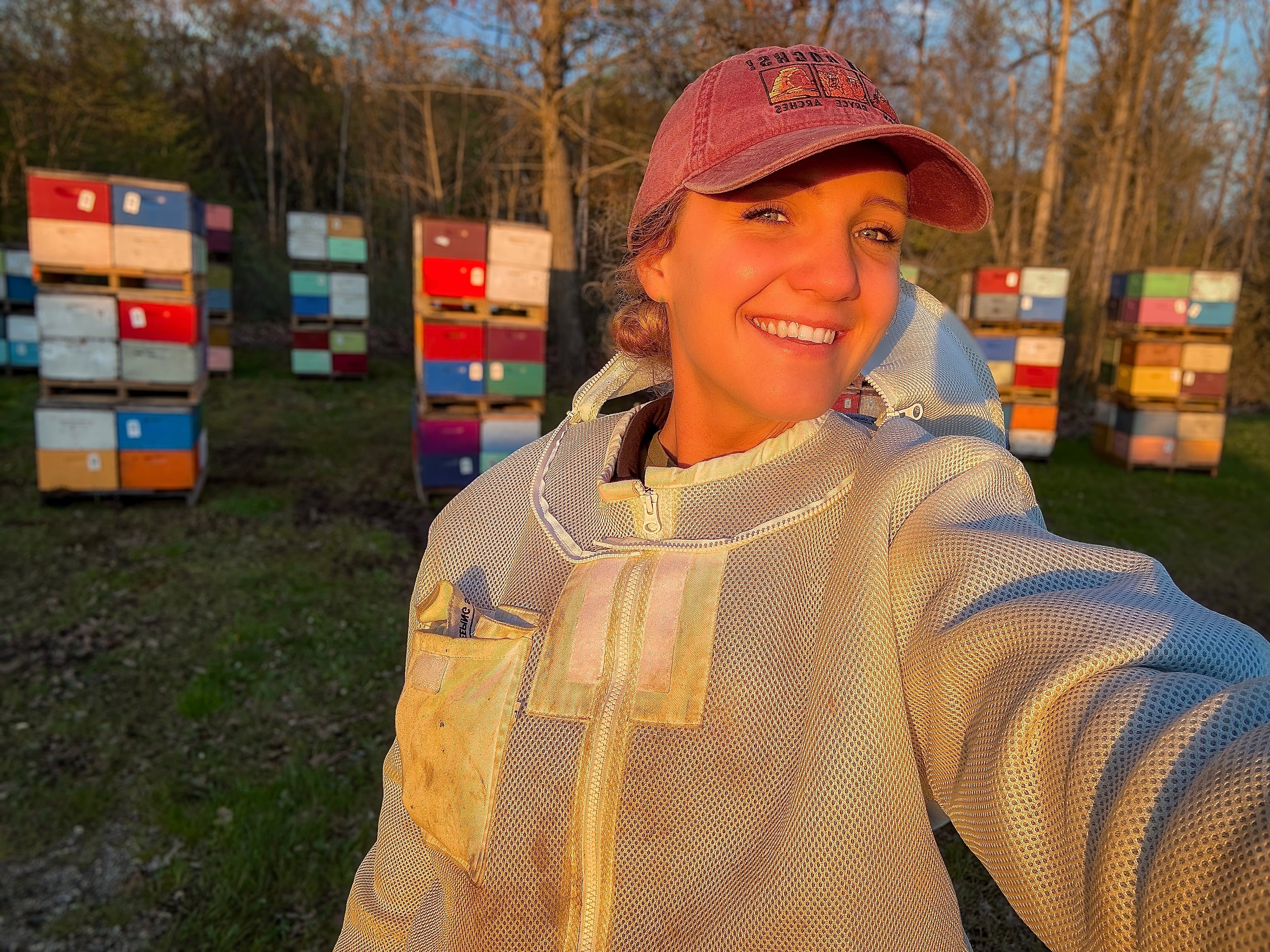 Emily from BeeFit Beekeeping smiling in a bee suit in front of stacks of honeybee hives